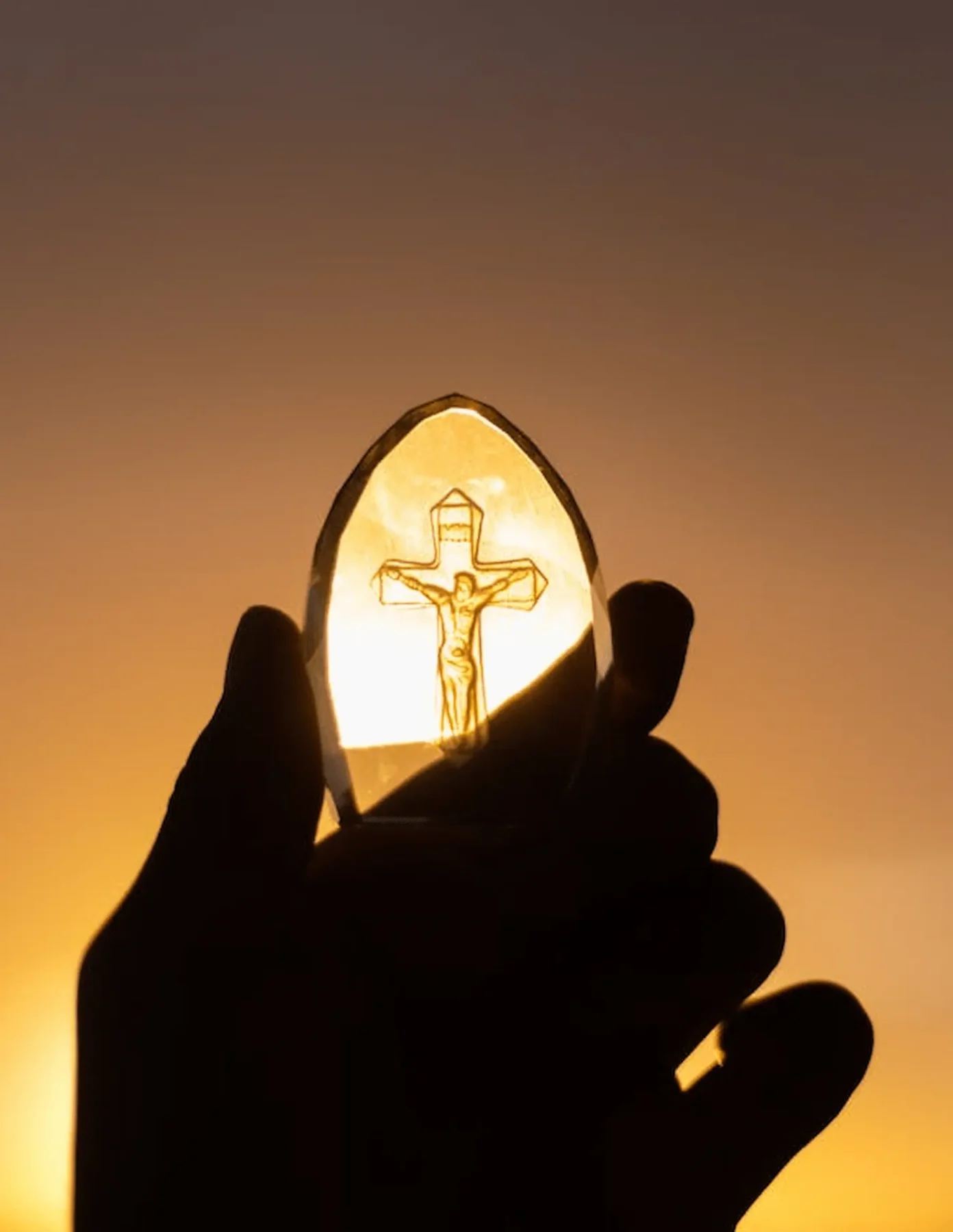 unas manos frente al sol aguantando una piedra con la imagen de Jesús en la cruz