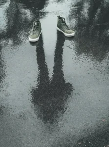 Unos zapatos bajo la lluvia y en el reflejo de la brea una persona