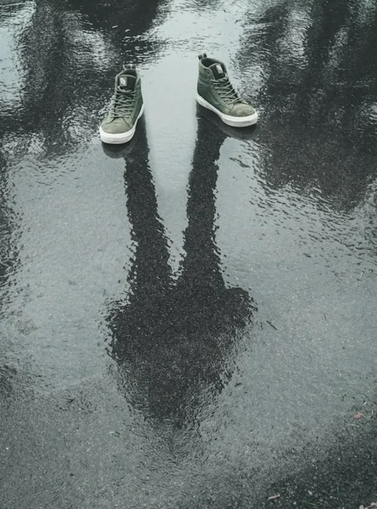 Unos zapatos bajo la lluvia y en el reflejo de la brea una persona