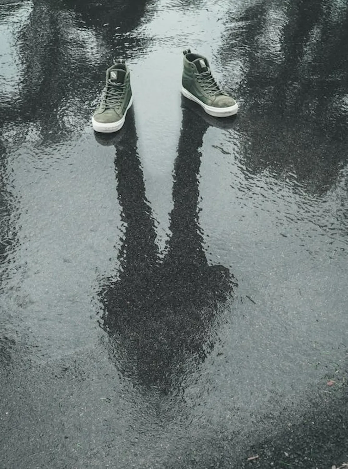 Unos zapatos bajo la lluvia y en el reflejo de la brea una persona