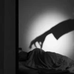 la sombra de una mano sobre la persona que está acostada en su cama y escondida bajo sus sabanas