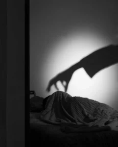 la sombra de una mano sobre la persona que está acostada en su cama y escondida bajo sus sabanas